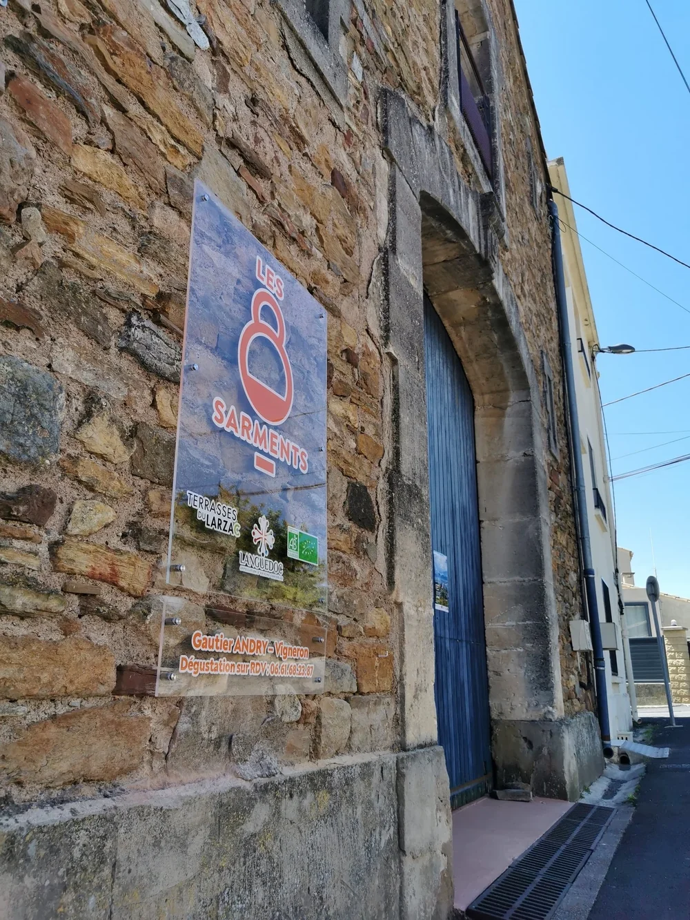 Façade en pierre de la cave avec l'enseigne 'Les 8 Sarments' (logos Terrasses du Larzac, bio, mention Gautier Andry vigneron / dégustation) et porte voûtée bleue. Identifie clairement le caveau.