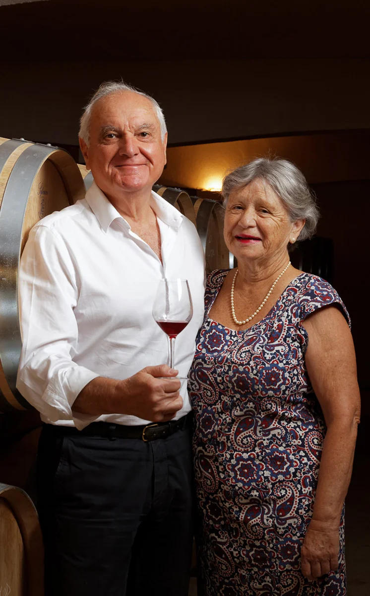 Portrait de couple Jean-Pierre et Maryse Rambier devant les barriques du chai, lui en chemise blanche avec un verre de rouge, elle en robe a motifs. Studio/lumiere soignee, tres digne et authentique.