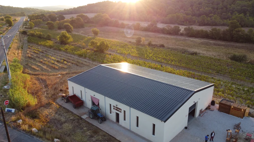 Magnifique vue aerienne (drone) du chai des Remparts de Cuculles a l'heure doree : batiment blanc a toit gris portant le nom du domaine, entoure de vignes et de collines de garrigue, soleil rasant. Hero shot ideal.