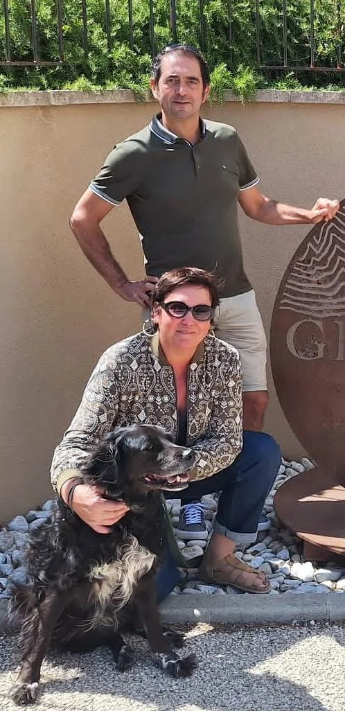 Portrait du couple fondateur Eric Michel (debout) et Marie-Luce Arnaud (accroupie) avec leur chien noir, posant devant l'entree du domaine pres d'une sculpture en acier. LA photo des vignerons, chaleureuse et authentique. Image de confiance ideale en page d'accueil ou page 'le domaine'.