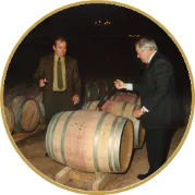 Photo d'archive : deux hommes en costume inspectant des barriques dans une cave ancienne, médaillon rond doré. Évoque l'histoire et le savoir-faire du chai.