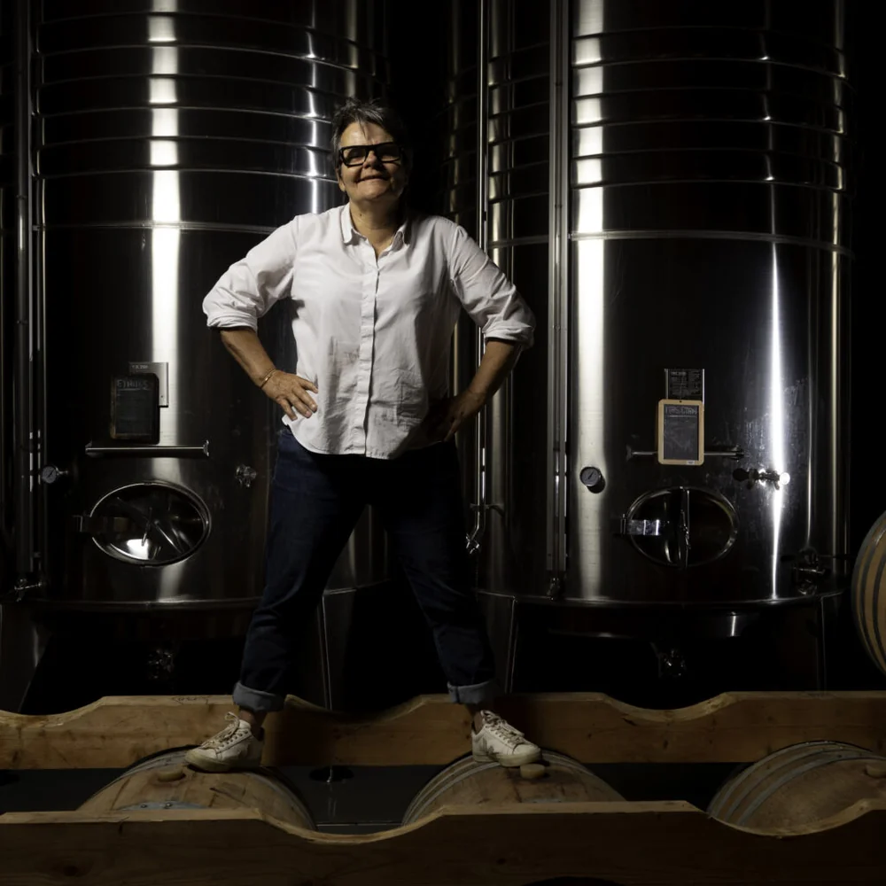 Portrait de Brigitte Jeanjean (vigneronne, 6e génération) debout dans le chai entre les cuves inox et les barriques, lumière dramatique. Photo professionnelle très valorisante, incarne l'humain et le savoir-faire.