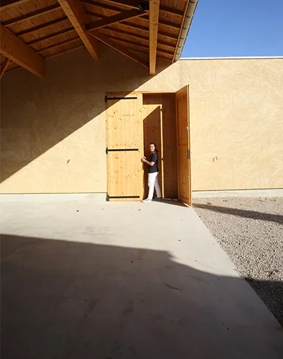 Entrée du chai contemporain : cour bétonnée, façade enduite ocre clair, grande porte en bois clair sous une charpente, une personne (Isabelle) se tenant dans l'embrasure pour donner l'échelle. Montre l'architecture épurée du domaine. Résolution native modeste (409x518), à réserver à un format réduit