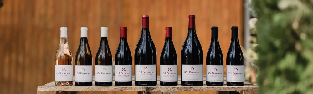 Alignement lifestyle de la gamme complète (rosé, blancs et rouges) posée sur une caisse en bois en extérieur, fond de bardage bois et végétation floue. Beau packshot d'ambiance valorisant l'ensemble de la collection (et non une étiquette plate).