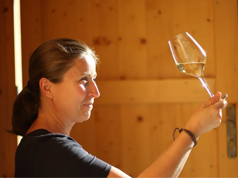 Portrait lumineux de la vigneronne (Isabelle Goumard) de profil, examinant un verre de vin blanc à contre-jour devant une cloison de bois clair du chai. Excellente photo : visage net, lumière chaude, geste de dégustation authentique. Image humaine la plus forte du domaine, idéale pour une page d'acc