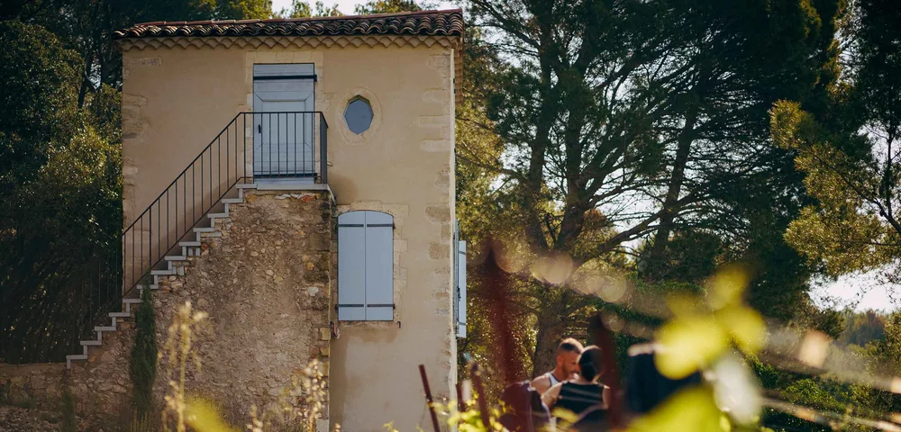 Facade du mas aux volets bleus avec son escalier exterieur en pierre ; un couple lit assis au premier plan parmi la vigne. Architecture du domaine avec une touche humaine, lumiere chaude.