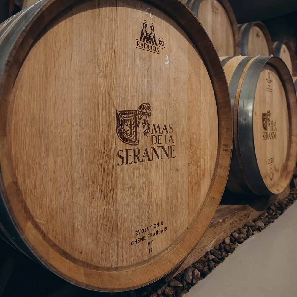 Barriques de chene marquees au fer 'Mas de la Seranne' alignees dans le chai, sur lit de galets. Belle photo de cave montrant le monogramme du domaine.