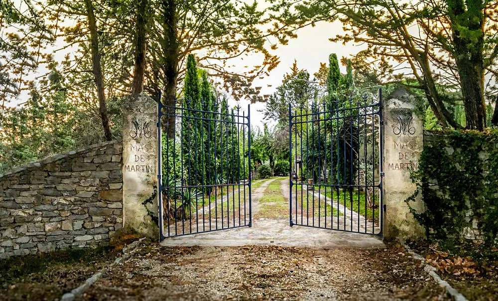 Le portail en fer forge a l'entree du domaine, encadre de piliers en pierre portant le monogramme 'Mas de Martin', allee bordee de cypres au coucher du soleil. Entree elegante et identitaire.
