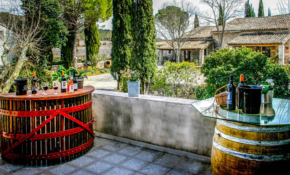 Mise en situation des bouteilles et verres sur des tonneaux/comptoir en bois, terrasse surplombant le mas et les vignes. Packshot lifestyle convivial dans le decor du domaine.