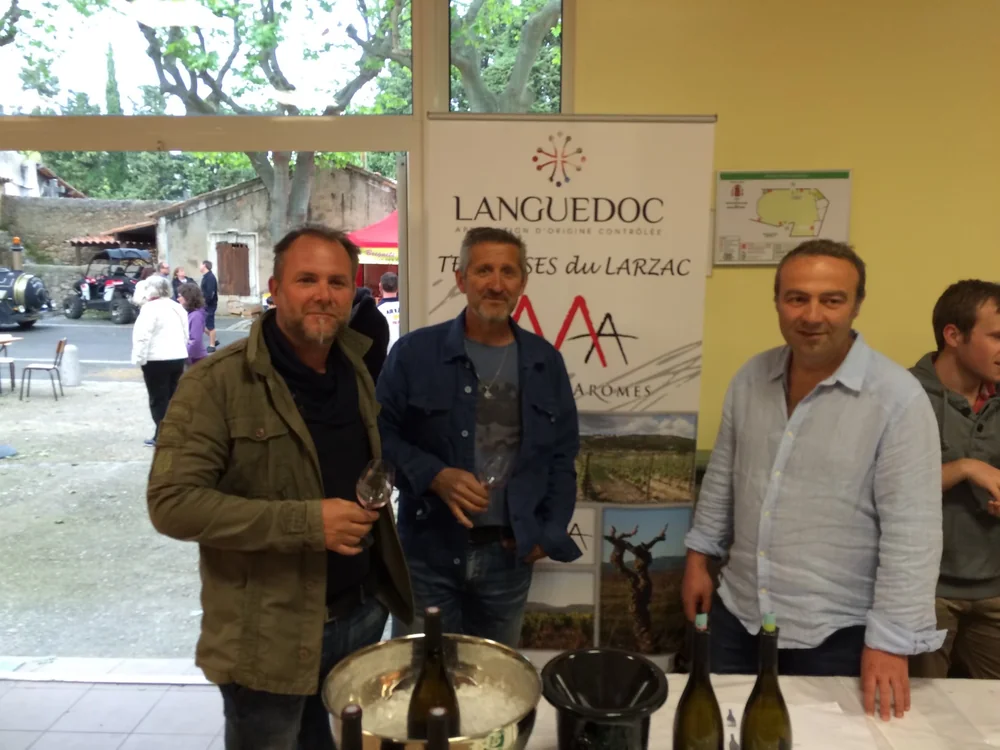 Les trois fondateurs/associes du domaine (Jean-Roch Cavalier, Didier Crezegut, Martial Garrigos), verre a la main, lors d'une degustation/salon, devant le kakemono Mas des Aromes (Languedoc / Terrasses du Larzac). Bouteilles et seau a glace au premier plan. Photo authentique, l'humain qui cree la co