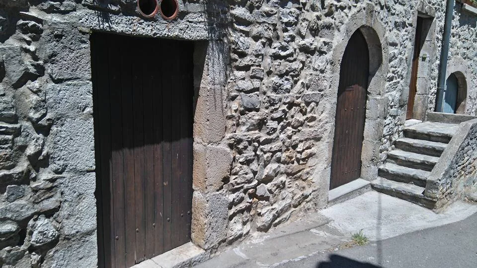 Facade en pierre du batiment du domaine dans le village (Viols-le-Fort) : murs de pierre seche, portes en bois ancien du cuvier (ancien fournil du XVIIe siecle), arche en pierre et escalier. Architecture languedocienne authentique, illustre l'ancrage patrimonial du domaine.